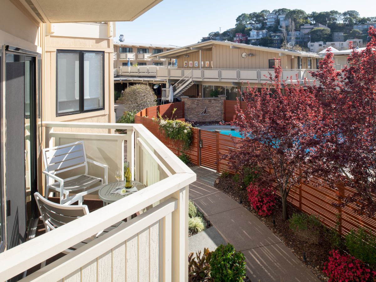 A balcony with white chairs and a small table overlooks a walkway with red trees and a pool, with buildings and a hill in the background.