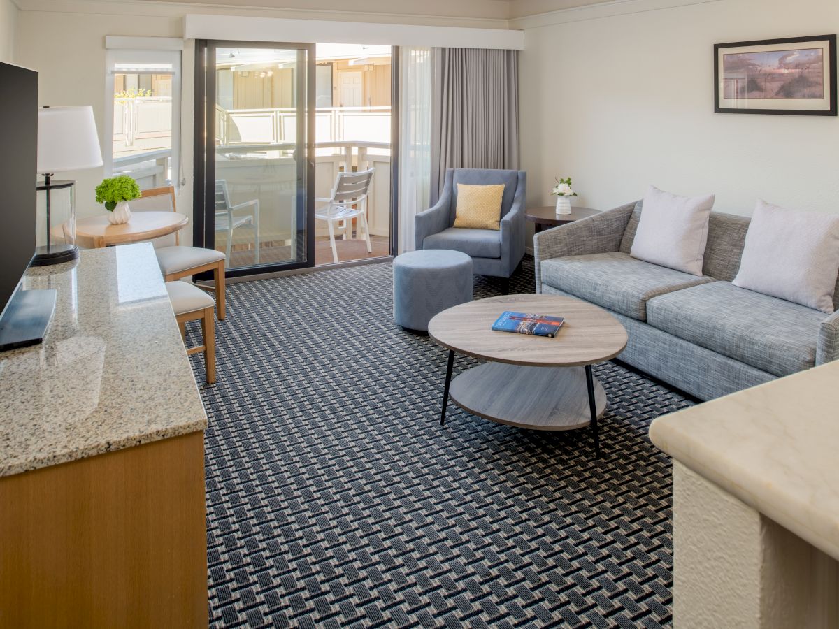 A modern living room features a gray sofa, coffee table, flat-screen TV, and an open balcony with chairs. The room is carpeted and well-lit.