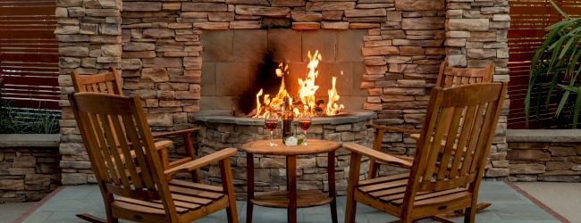 Two wooden rocking chairs face a burning outdoor fireplace, with a small wooden table in between holding a couple of drinks and a candle.