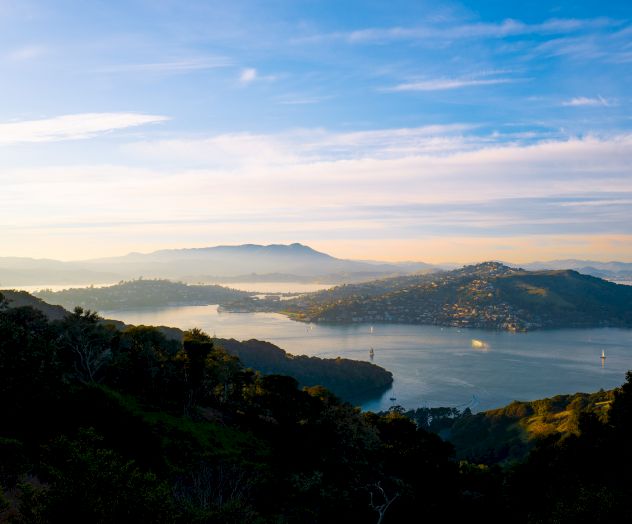 The image shows a scenic view of a hilly landscape with water bodies, covered in mist and surrounded by greenery under a blue sky with clouds.