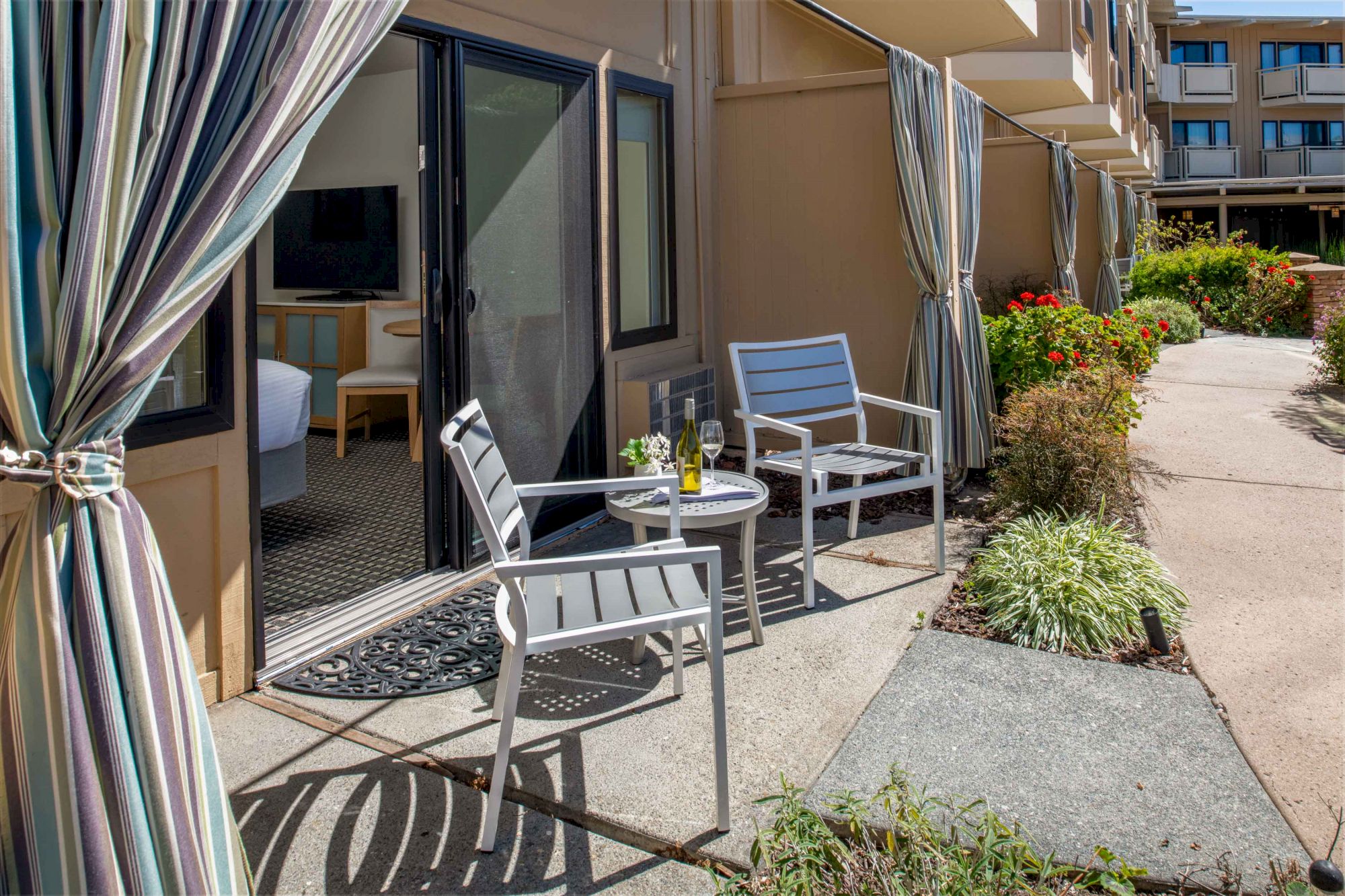 A patio with a small round table, two chairs, a bottle of wine, and glasses, next to a sliding door with curtains and a garden area.