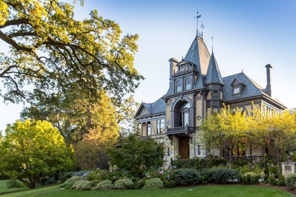 This image showcases a large, elegant mansion with intricate architecture, surrounded by lush greenery and trees, under a clear blue sky.