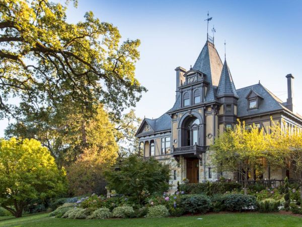 This image showcases a large, elegant mansion with intricate architecture, surrounded by lush greenery and trees, under a clear blue sky.
