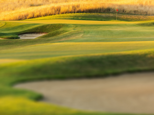 A lush golf course is shown with well-maintained greens, sand bunkers, and a distant red flag indicating the hole.