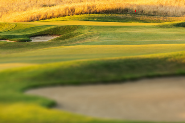 A lush golf course is shown with well-maintained greens, sand bunkers, and a distant red flag indicating the hole.