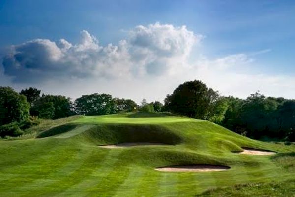 A lush green golf course with sand bunkers, rolling hills, and trees under a partly cloudy blue sky is depicted in the image.