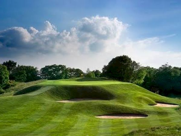 A lush green golf course with sand bunkers, rolling hills, and trees under a partly cloudy blue sky is depicted in the image.