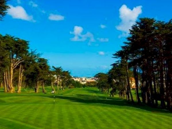 The image shows a green and well-maintained golf course surrounded by trees, with a blue sky and some buildings visible in the background.