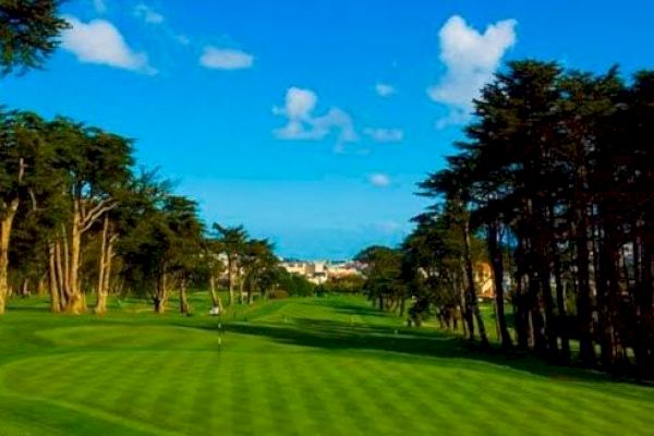 The image shows a green and well-maintained golf course surrounded by trees, with a blue sky and some buildings visible in the background.