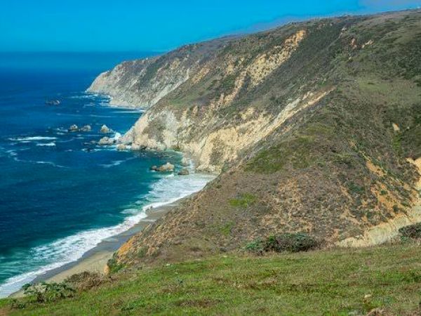A rugged coastline with steep cliffs, surrounded by blue ocean waves, under a clear blue sky, with patches of green grass on the hillsides.