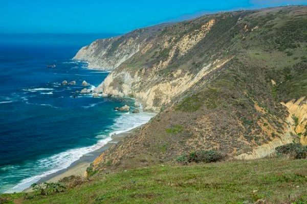 A rugged coastline with steep cliffs, surrounded by blue ocean waves, under a clear blue sky, with patches of green grass on the hillsides.