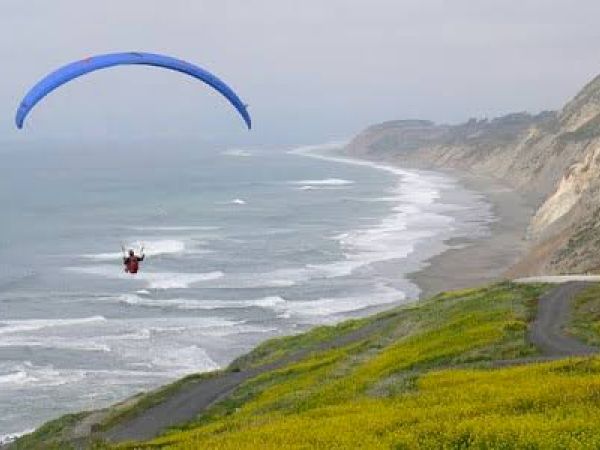 A person is paragliding over a coastal landscape with cliffs, a winding road, and yellow-green vegetation by the ocean.