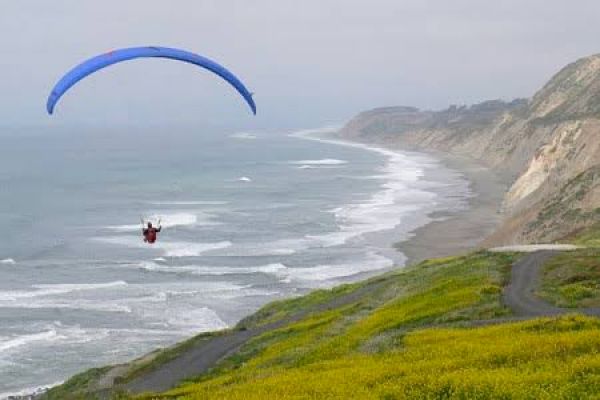 A person is paragliding over a coastal landscape with cliffs, a winding road, and yellow-green vegetation by the ocean.