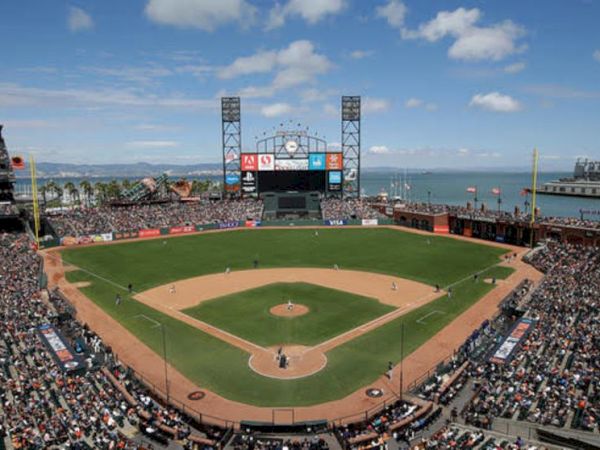 A packed baseball stadium with a game in progress, situated near a body of water under a partly cloudy sky with a large scoreboard in the outfield.