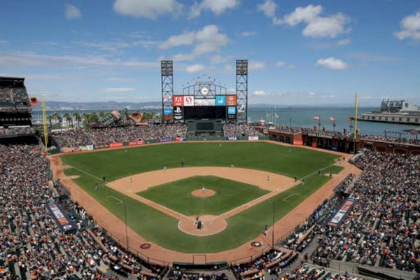 A packed baseball stadium with a game in progress, situated near a body of water under a partly cloudy sky with a large scoreboard in the outfield.