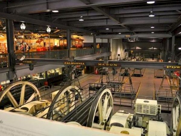 The image shows an interior view of a cable car museum with displays, historical cable cars, and large machinery parts.