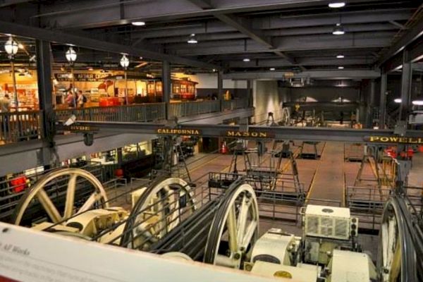 The image shows an interior view of a cable car museum with displays, historical cable cars, and large machinery parts.