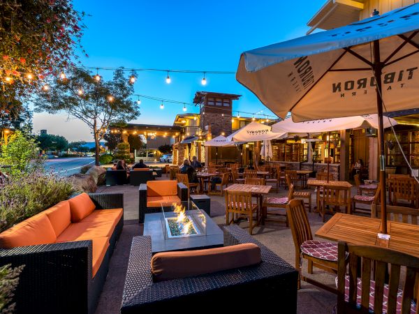 An outdoor restaurant patio at dusk with string lights, tables, chairs, a fire pit, and large umbrellas creating a cozy atmosphere.