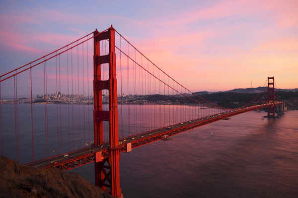 A red suspension bridge spans a large body of water at sunset, with a city skyline in the background under a colorful sky.