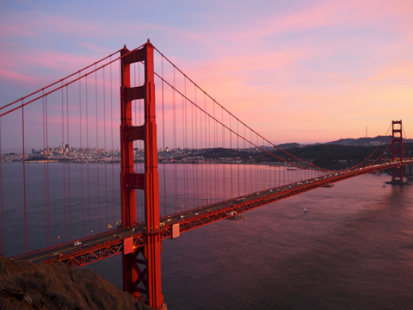 A red suspension bridge spans a large body of water at sunset, with a city skyline in the background under a colorful sky.