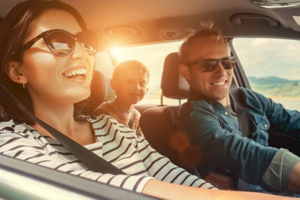 A family is enjoying a car ride with the sun shining; all are wearing sunglasses and seem content and happy while driving through a scenic route.