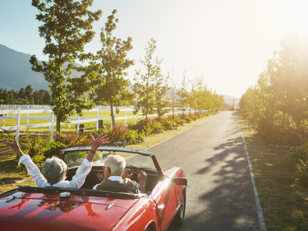 A couple is driving in a red convertible along a scenic road surrounded by trees, with one raising their arms in joy under the sunny sky.