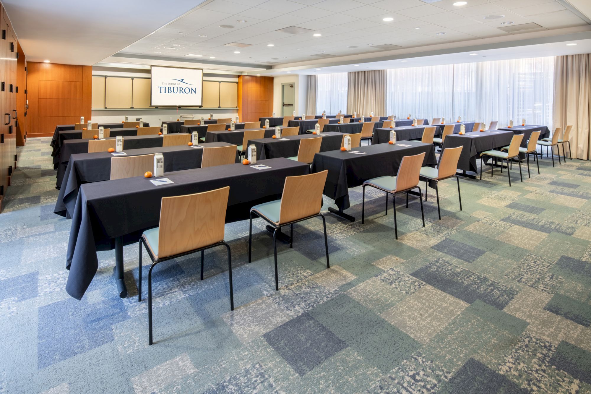 A conference room with arranged tables and chairs facing a screen, each table with water bottles and paper. The screen says 