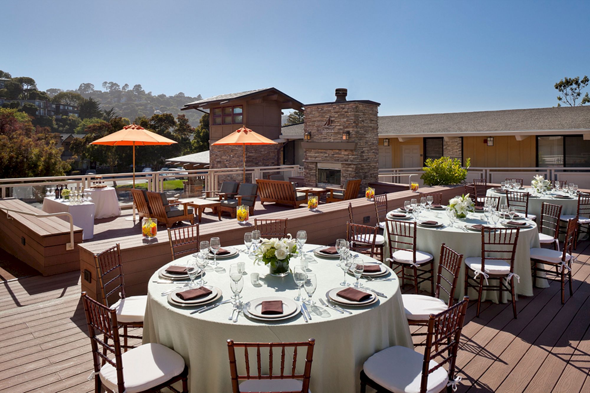 The image shows an outdoor terrace set up for an event with round tables, chairs, a stone fireplace, and orange umbrellas under a clear sky.