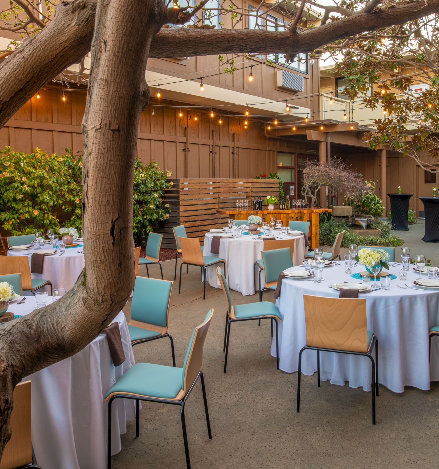 A beautifully set outdoor dining area with round tables, white tablecloths, and green chairs, surrounded by foliage and string lights for ambiance.