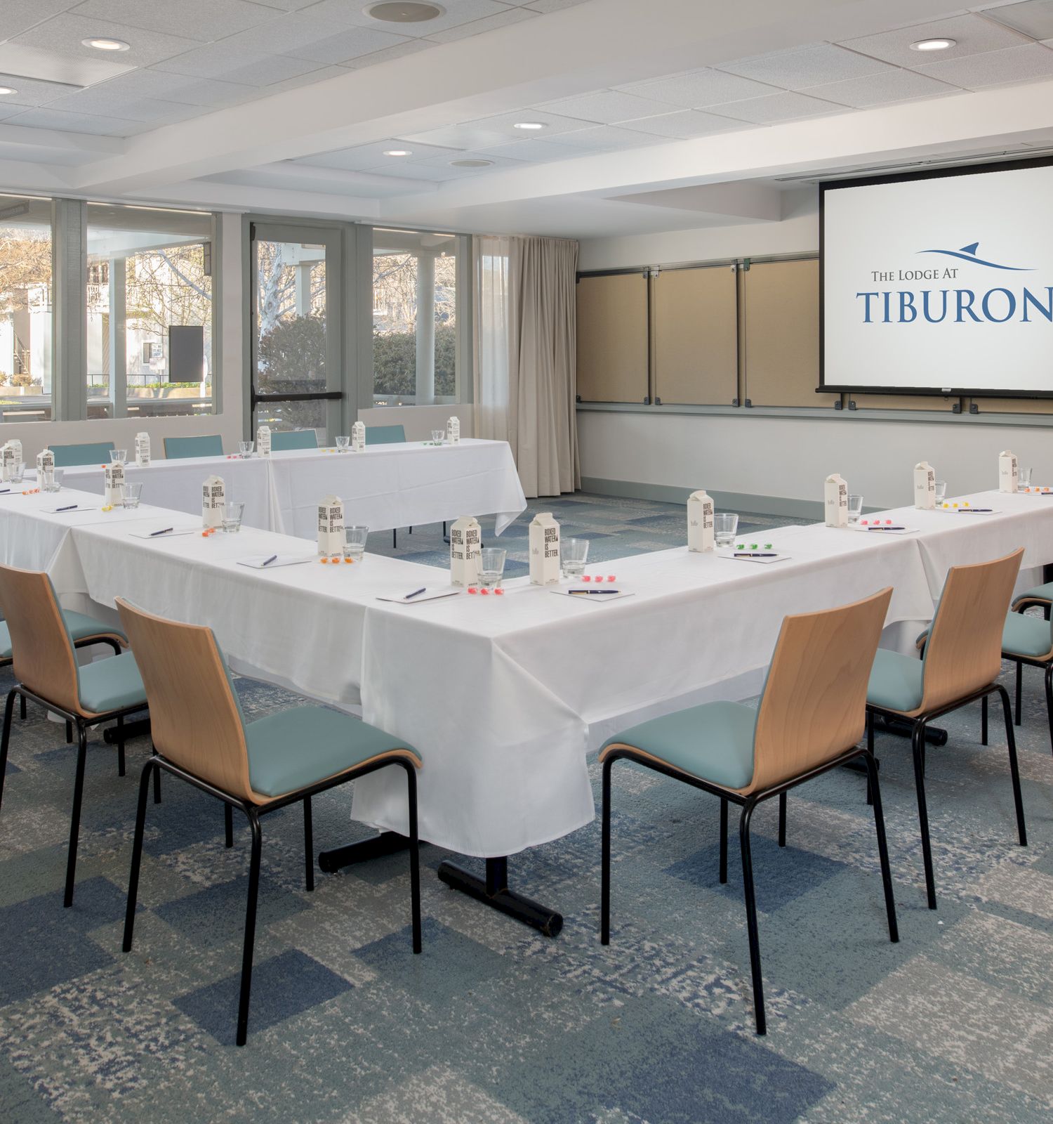 The image shows a conference room setup with a U-shaped table arrangement, chairs, a screen displaying 