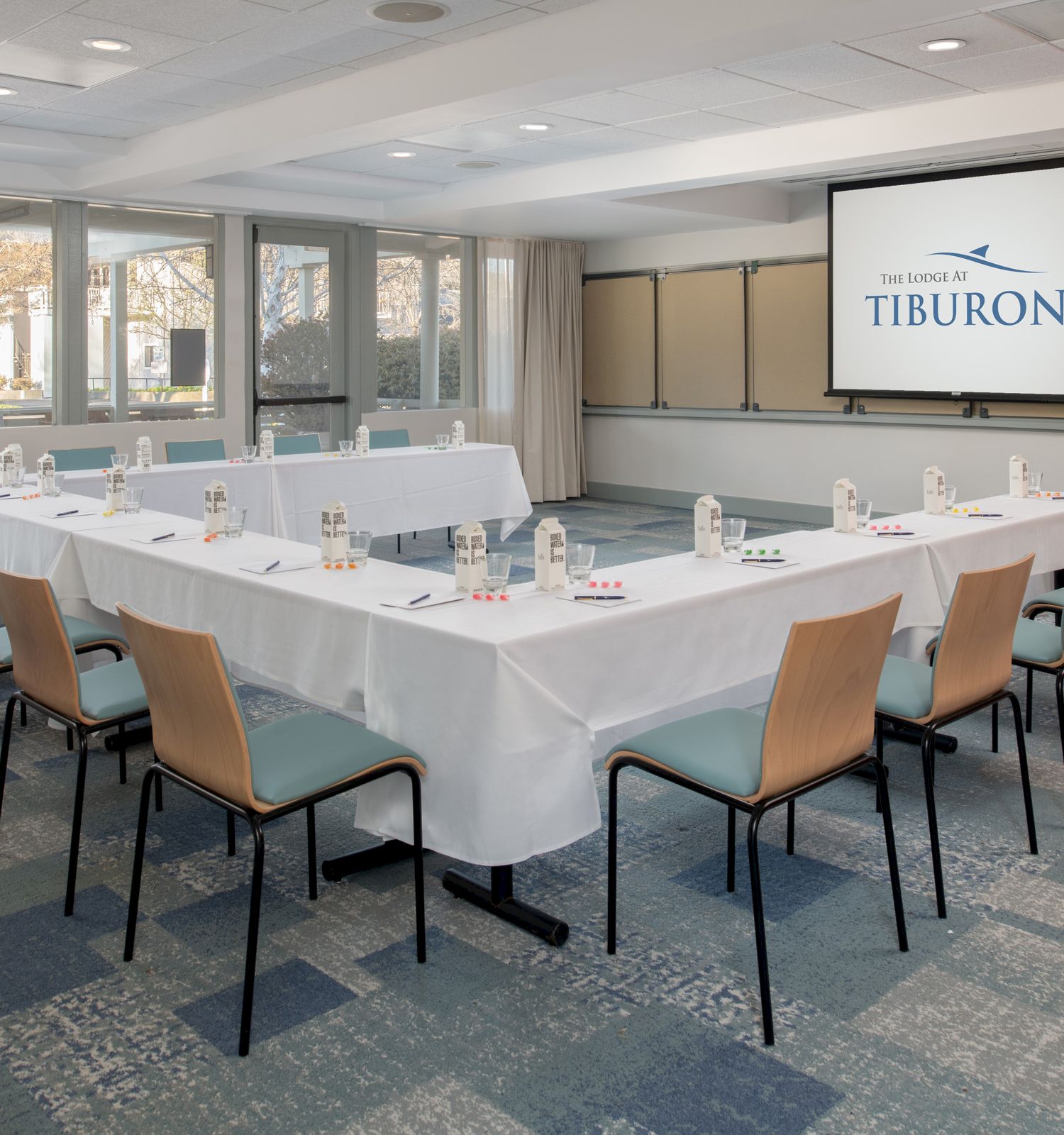 The image shows a conference room setup with a U-shaped table arrangement, chairs, a screen displaying "THE LODGE AT TIBURON," and multiple windows.