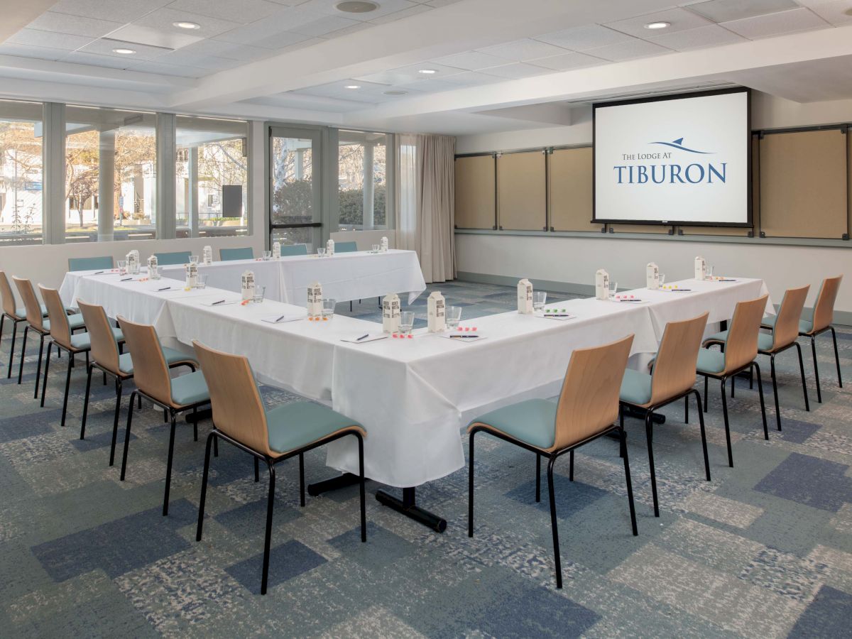 A conference room features a U-shaped table setup, chairs, water bottles, and a screen displaying "Tiburon." The room is bright, with large windows.