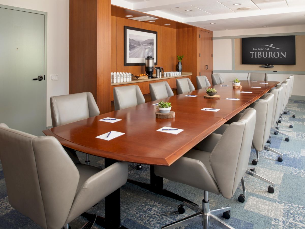 A conference room with a long wooden table, beige chairs, small plants, note pads, and a screen displaying “Tiburon” is shown.