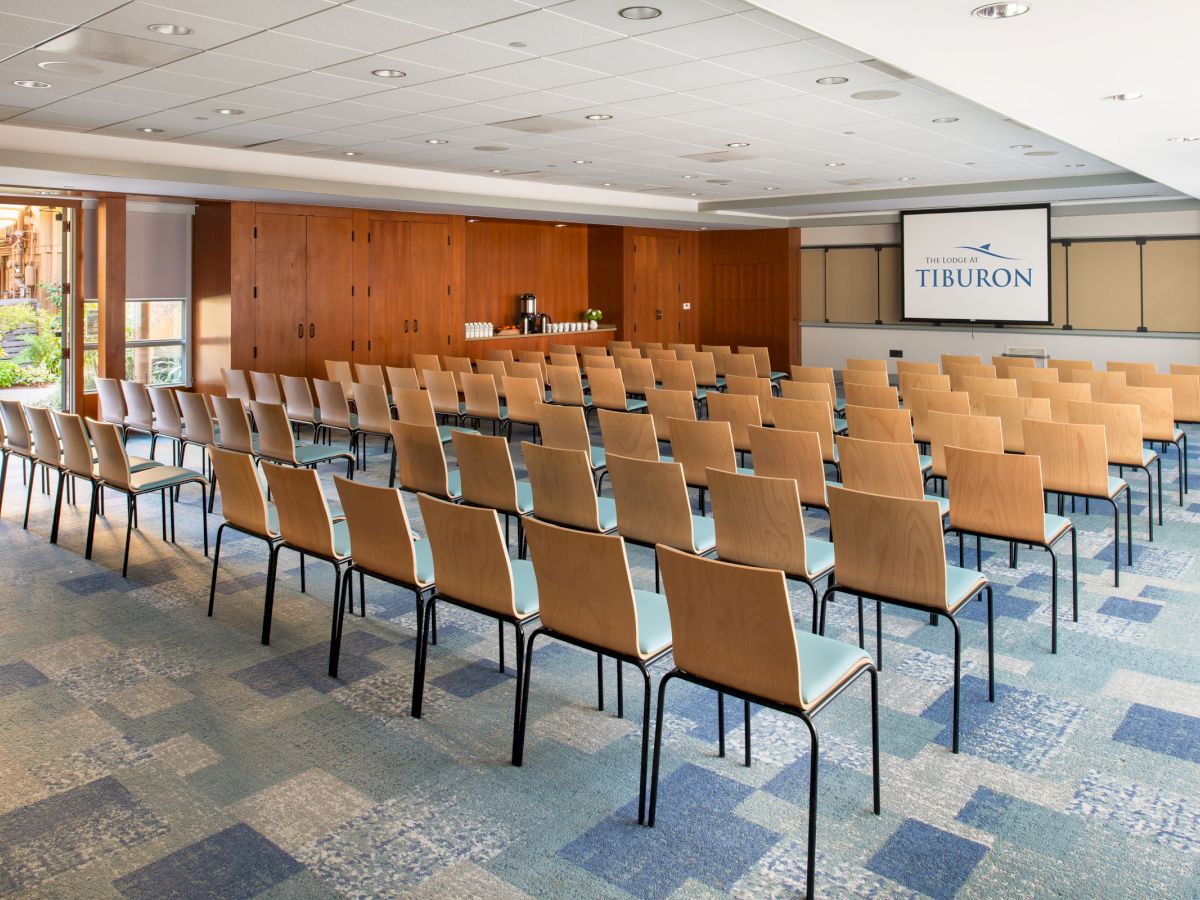 The image shows a conference room with rows of empty chairs facing a screen at the front displaying 