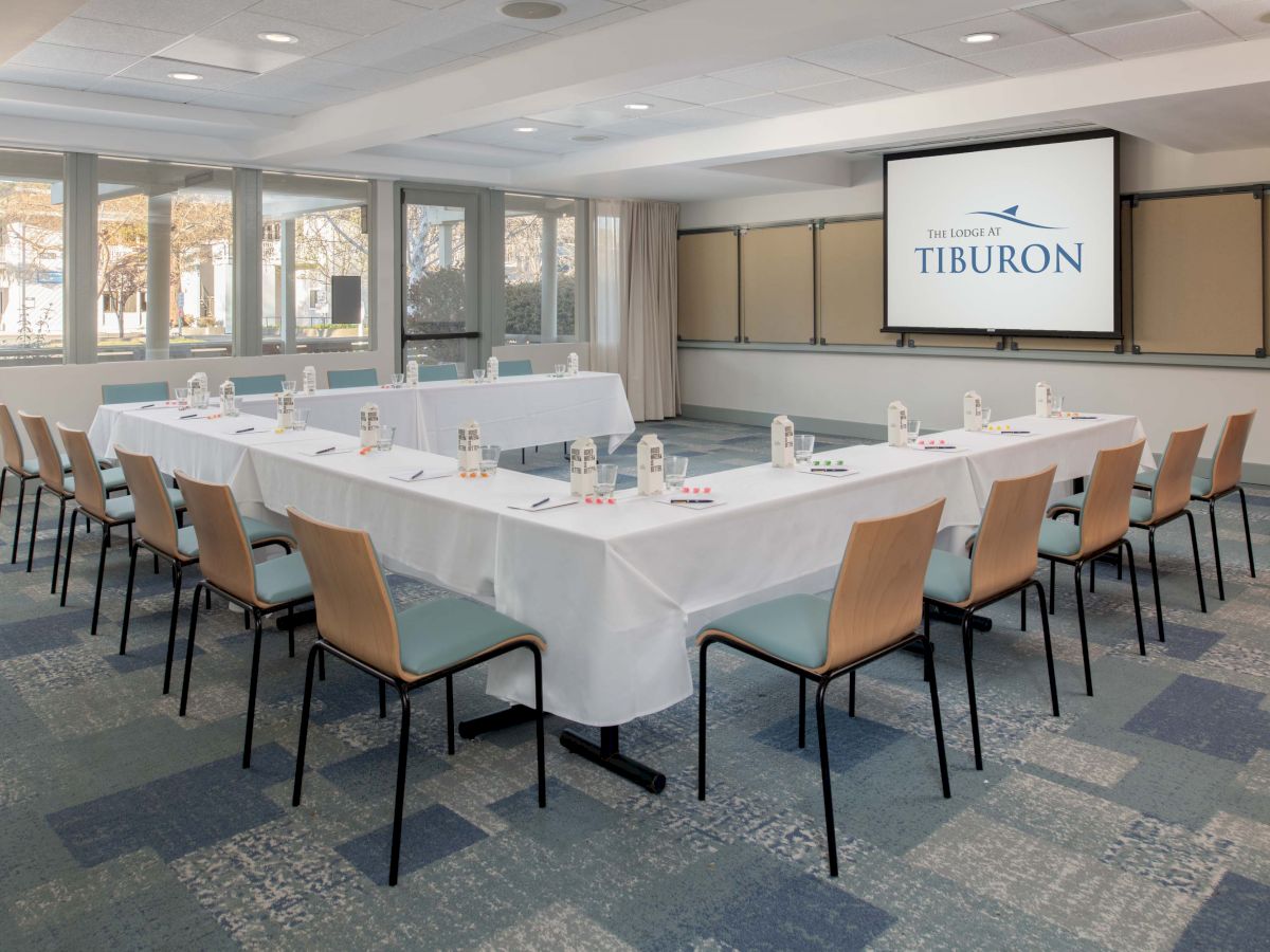 A conference room features a U-shaped table setup, chairs, water bottles, and a screen displaying 