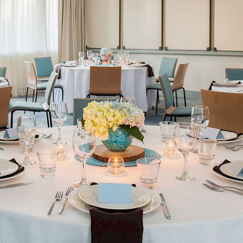 A beautifully set dining table with white cloths, flowers in glass vases, wine glasses, cutlery, and neatly arranged chairs in a bright, elegant room.