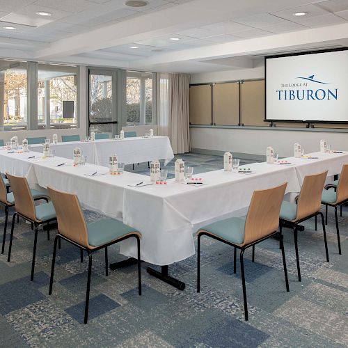 A conference room with a U-shaped table setup, chairs, notebooks, pens, and water bottles. There's a screen displaying 