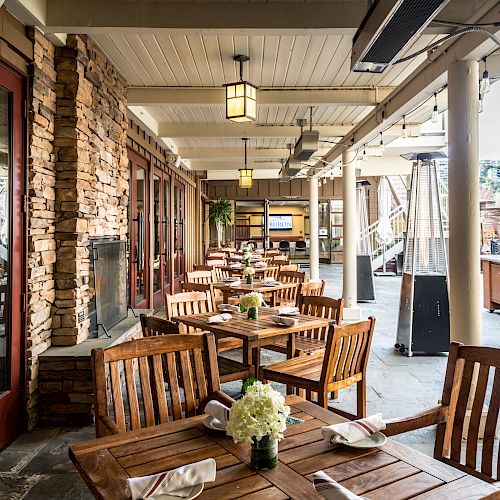 An outdoor restaurant seating area with wooden tables and chairs, napkins, and a centerpiece, under a covered patio with stone walls and ceiling lights.