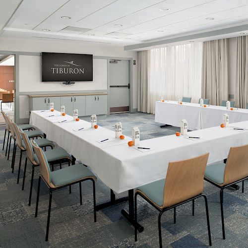 The image shows a conference room setup with a U-shaped table layout, chairs, water bottles, and notepads, facing a screen that displays 