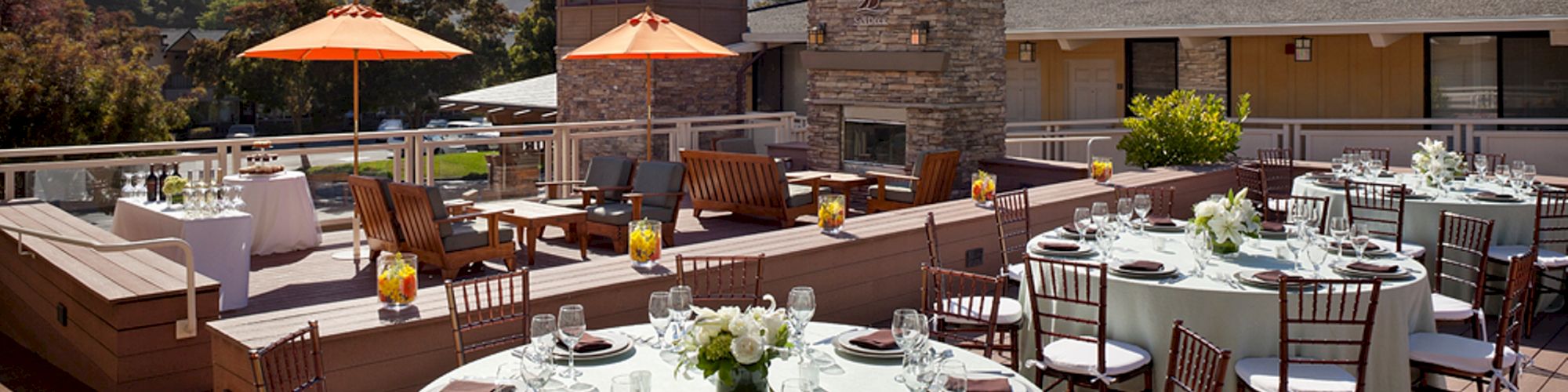 An outdoor terrace set up for an event with round tables, chairs, place settings, and orange umbrellas, overlooking a scenic landscape under a blue sky.