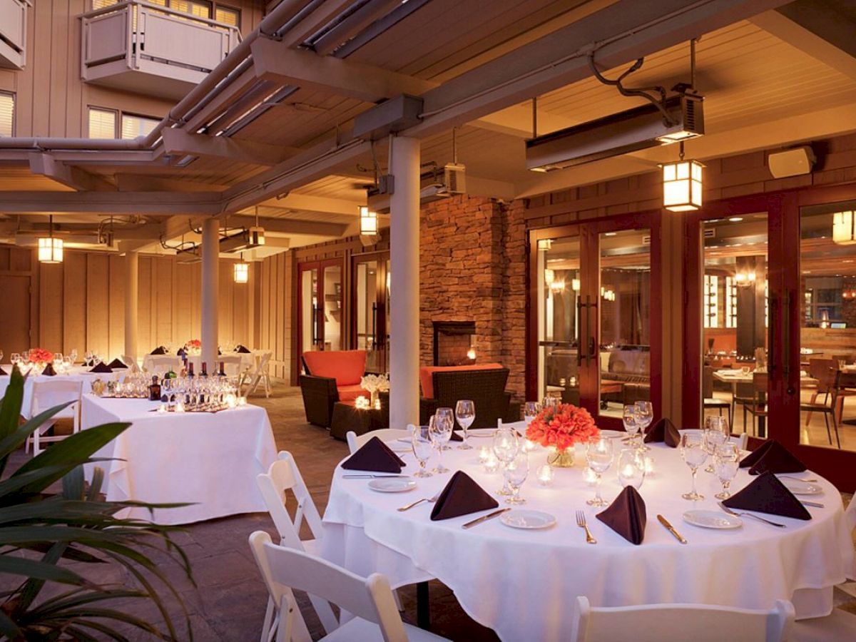 An outdoor dining area with round tables set with white tablecloths, black napkins, glassware, and floral centerpieces, near a cozy, lamp-lit patio.