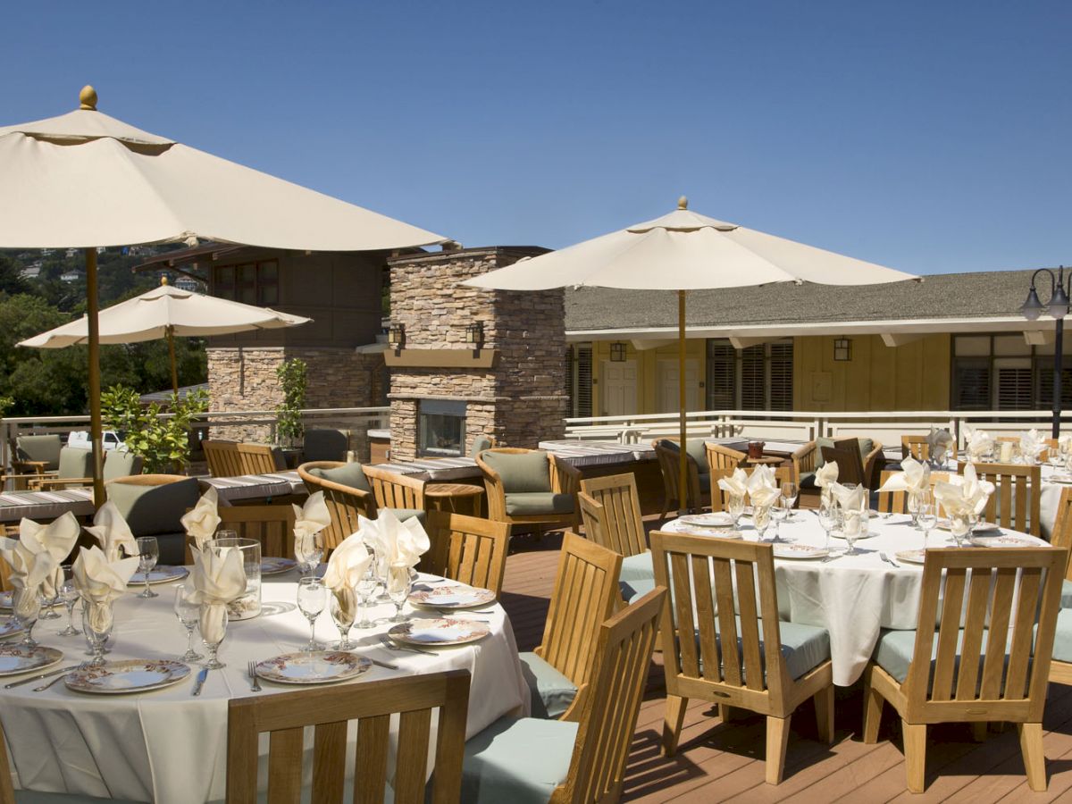 An outdoor patio with round tables set for dining, wooden chairs, white tablecloths, folded napkins, and large umbrellas providing shade.