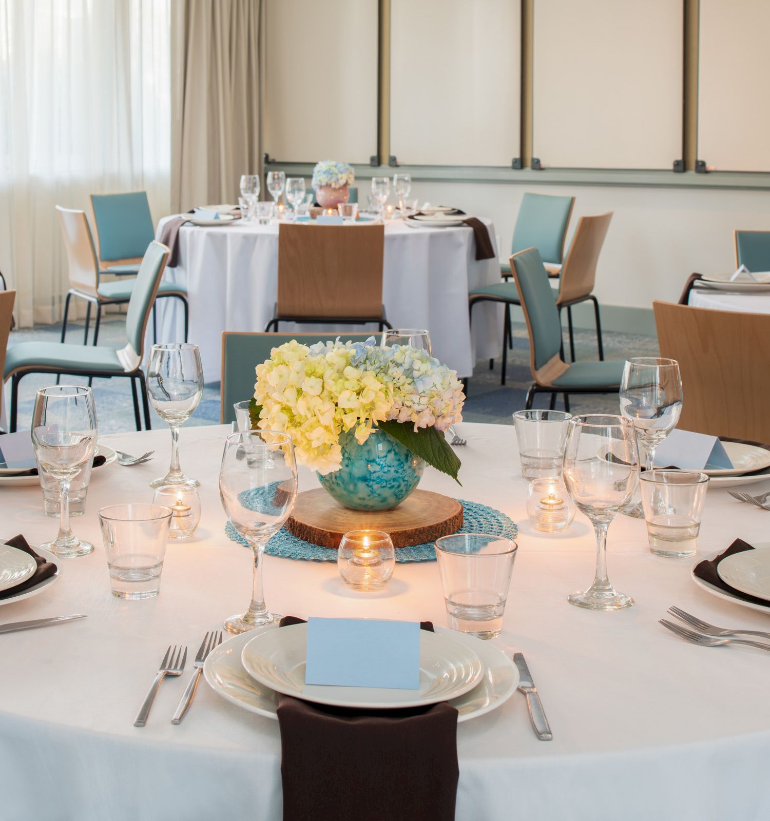 The image shows a banquet setup with round tables, white tablecloths, elegant place settings, and floral centerpieces in a bright room with chairs.