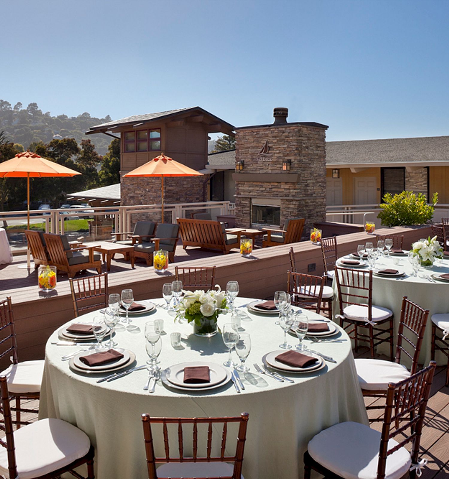 The image shows an outdoor patio setup for an event, with round tables, chairs, place settings, orange umbrellas, and a stone fireplace in the background.