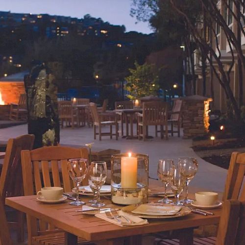 An outdoor dining area at dusk with wooden tables, chairs, and a lit candle centerpiece, surrounded by a warm, inviting atmosphere with a distant fire pit.