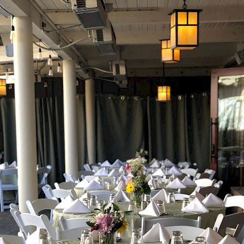 The image shows an indoor dining area with tables set for an event, decorated with flowers, folded napkins, and warm lighting, suggesting a formal gathering.