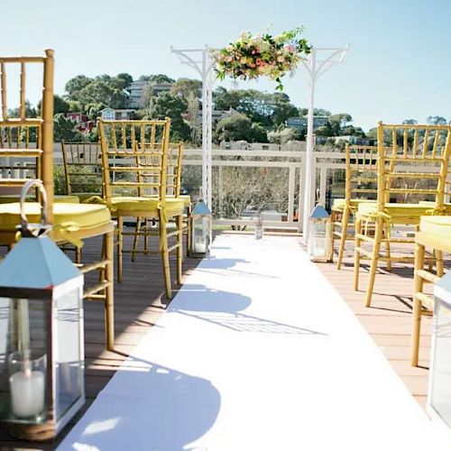 An outdoor wedding setup with yellow chairs, an aisle runner, lanterns with candles, and a floral arch. The background shows trees and a hill.