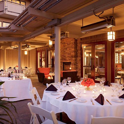 An outdoor dining area with round tables set with white tablecloths, black napkins, wine glasses, and a centerpiece of flowers, under warm lighting.