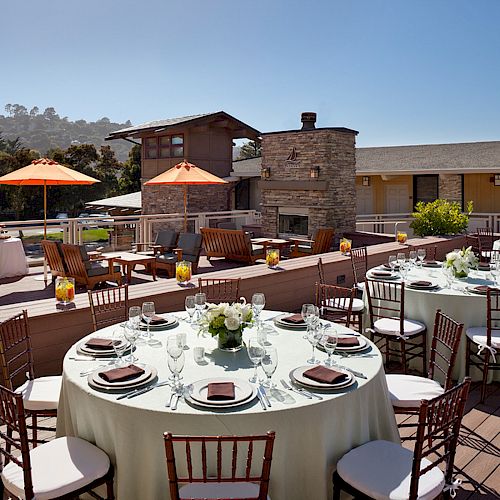 An outdoor patio setting with round tables arranged for a gathering, featuring parasols, a fireplace, and scenic views in the background.