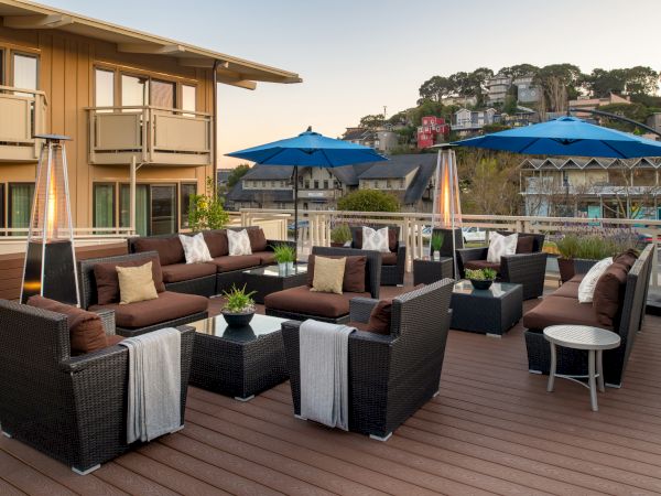 This image shows an outdoor patio area with wicker seating, cushions, blue umbrellas, and small tables, set against a backdrop of buildings and trees.